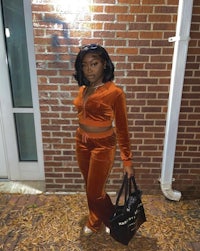 a woman in an orange velvet jumpsuit posing in front of a brick wall